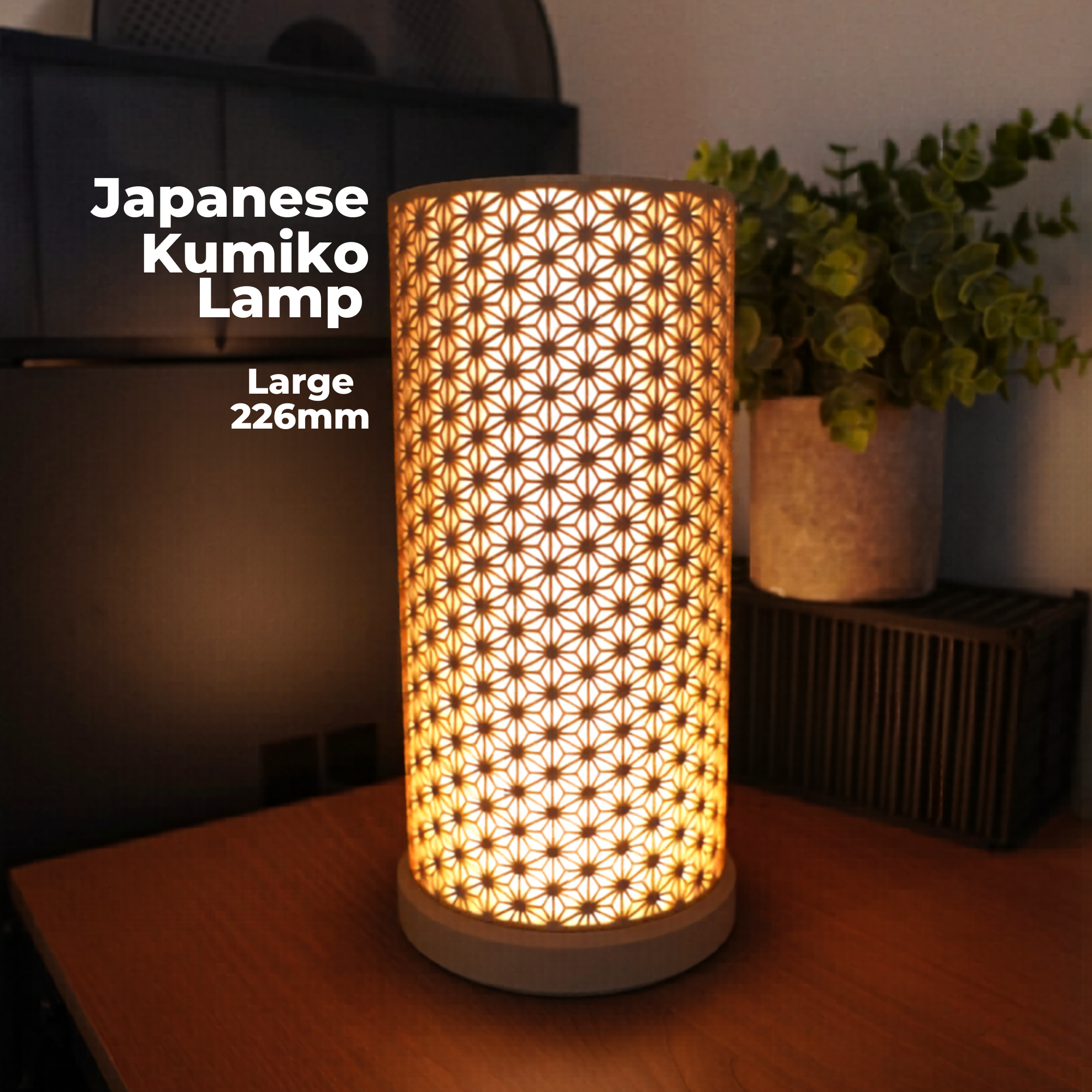 A lit cylindrical Japanese Kumiko lamp with a geometric star-pattern cutout shade, shown on a wooden table in a home setting with a potted plant in the background. White text on the left reads 'Japanese Kumiko Lamp' and 'Large 226mm'.