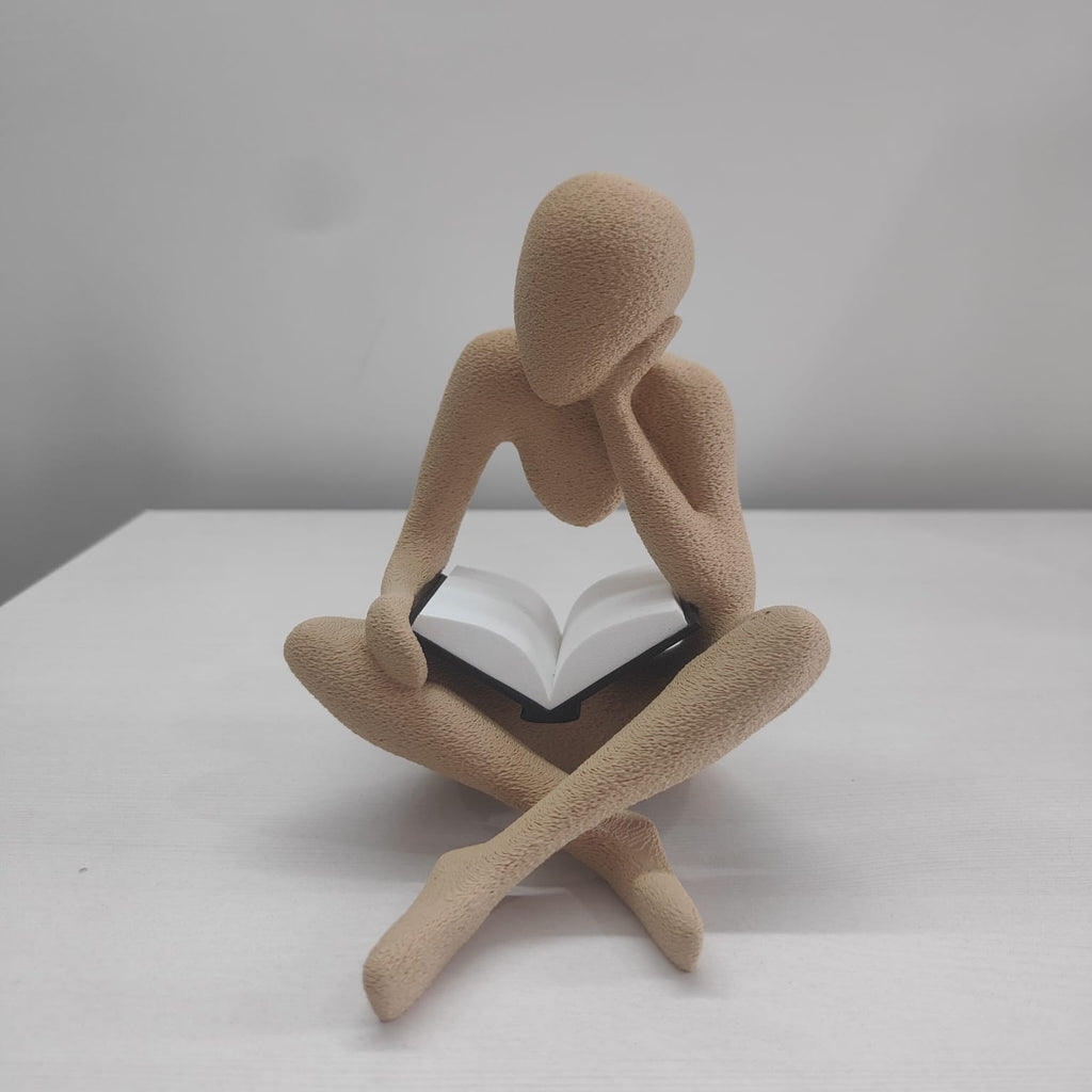 A minimalist beige figurative sculpture of a seated person holding an open book, displayed on a white surface against a light gray background.