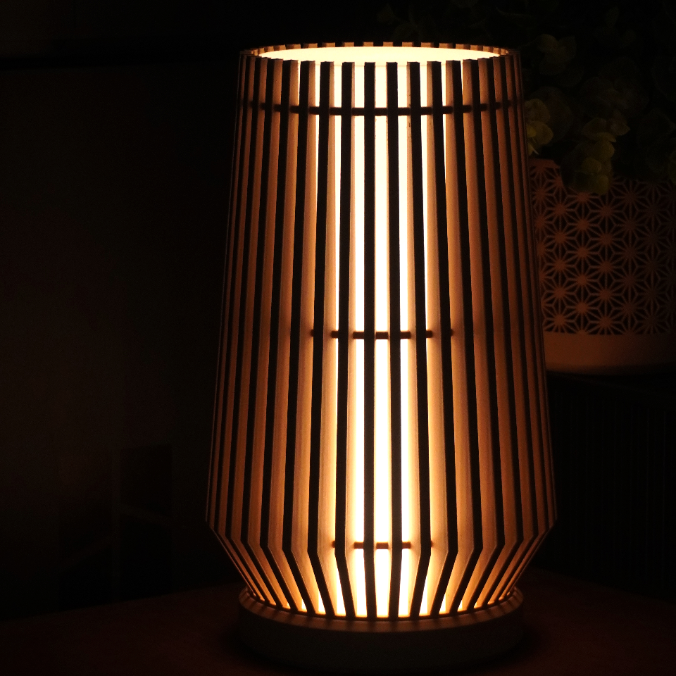 A modern bamboo table lamp with a ribbed cylindrical shade glowing with warm light, set on a dark wooden surface in a dimly lit room.