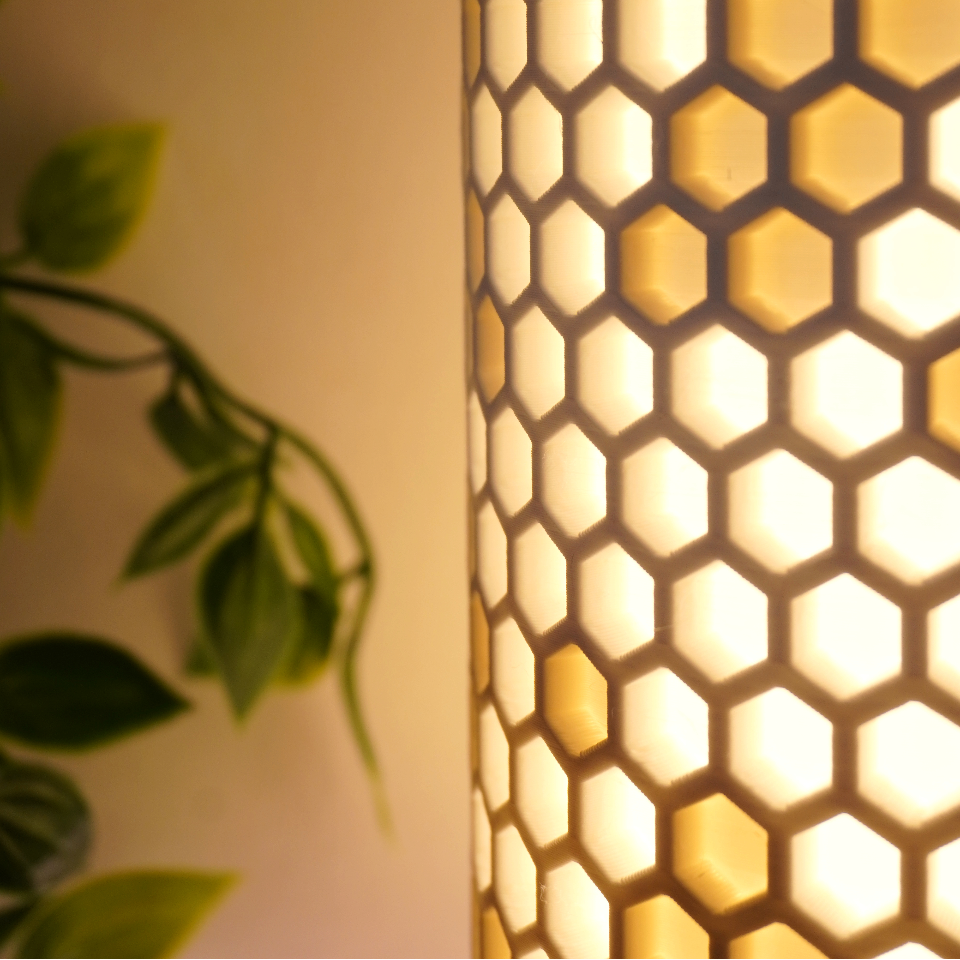 A close-up of a cylindrical lamp with a honeycomb pattern, glowing with warm yellow light against a beige wall with blurred green leaves on the left.