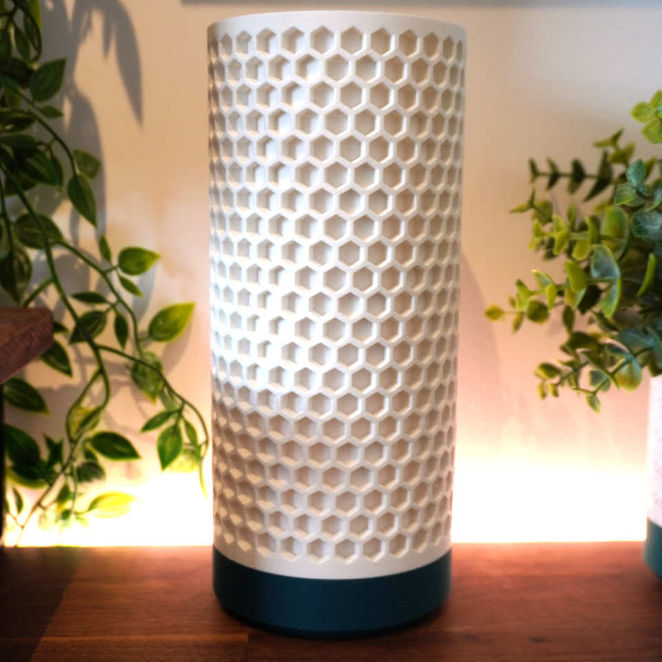 A modern cylindrical table lamp with a white honeycomb-textured shade and a dark teal base, sitting on a wooden surface with warm backlighting and plants on either side.