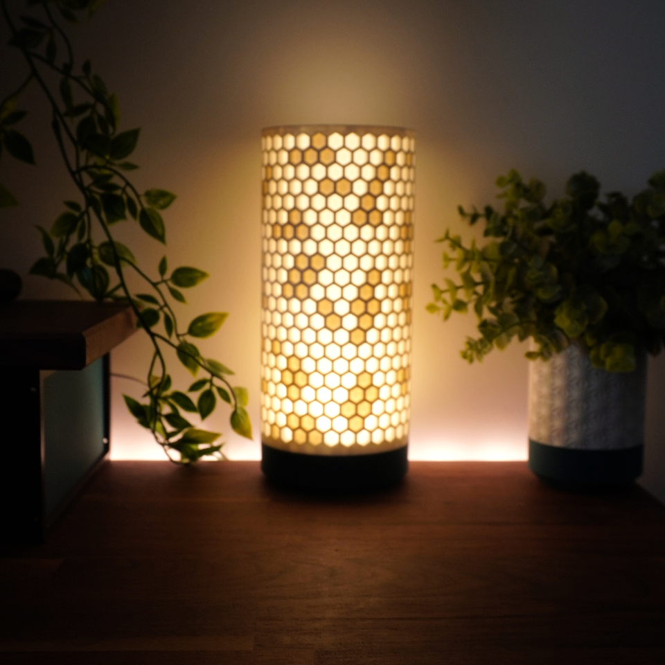 A glowing cylindrical table lamp with a honeycomb pattern shade and a black base, placed on a wooden surface between two potted plants in a dimly lit room.