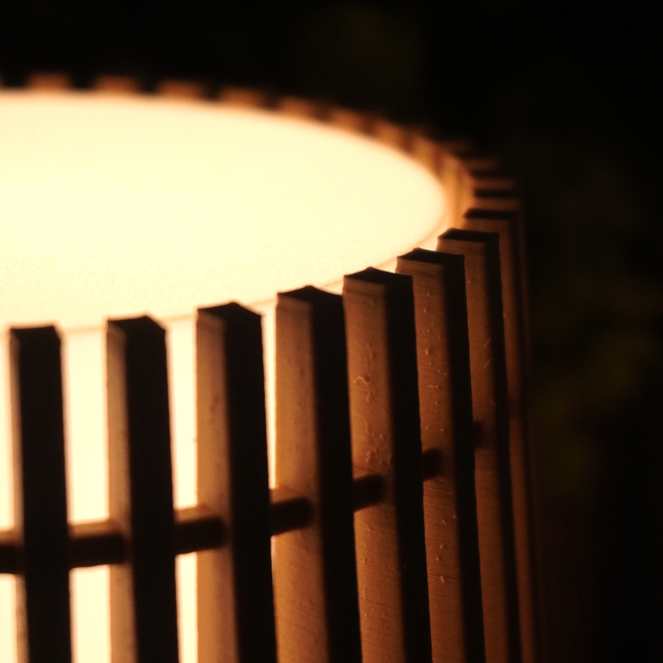 Close-up of a warm glowing cylindrical lamp or lantern with vertical slats, photographed in low light against a dark background.