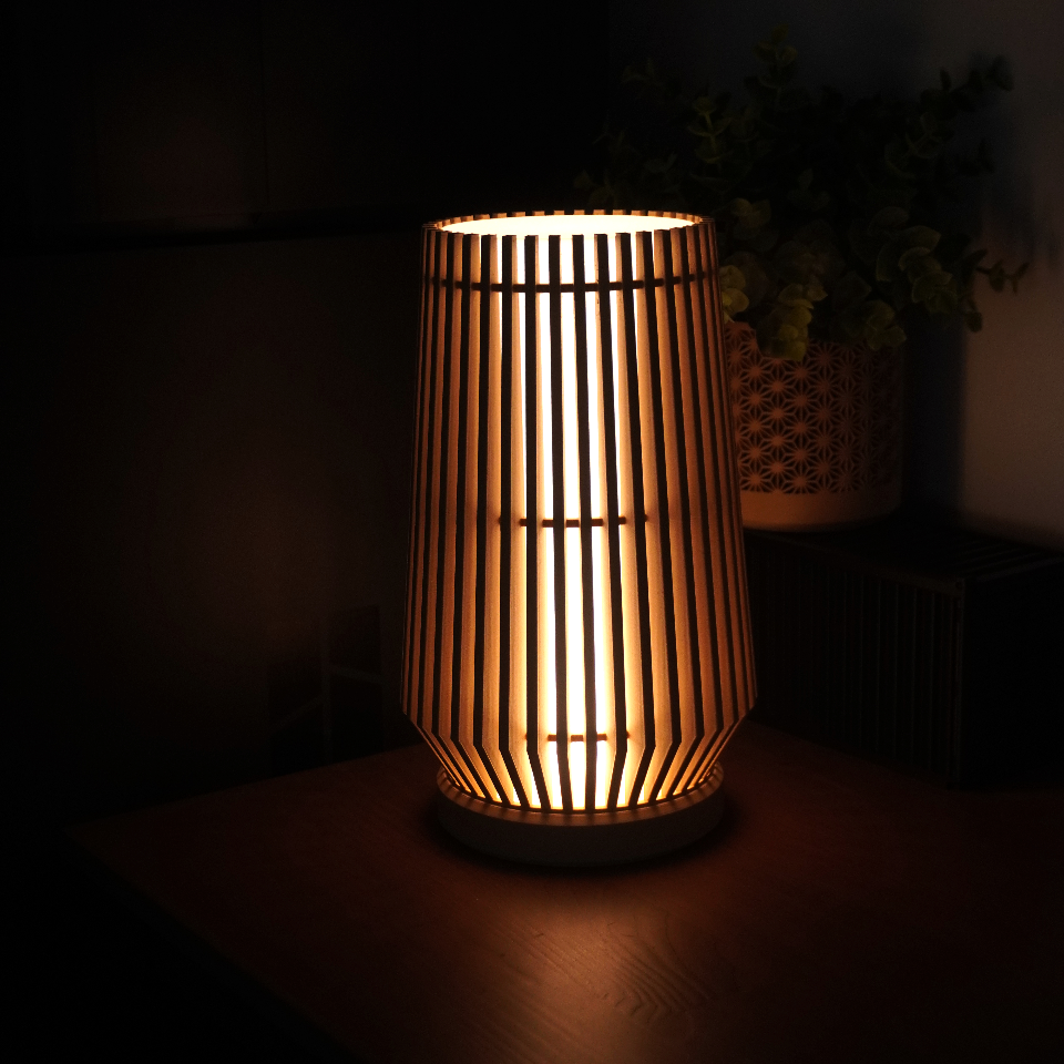 A warm glowing cylindrical table lamp with vertical slats, placed on a dark wooden surface in a dimly lit room with a plant in the background.