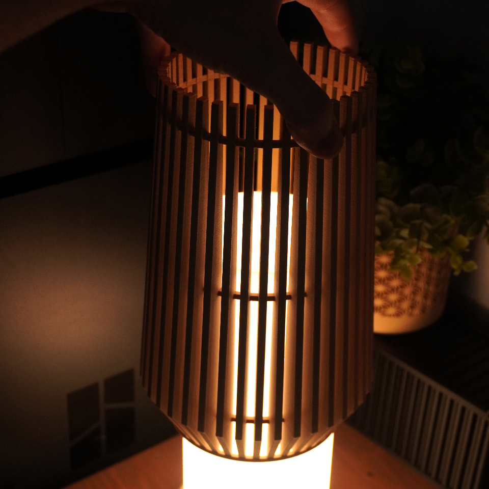 A bamboo-style table lamp with a cylindrical shade made of vertical slats, glowing warmly from a bright white light inside, shown on a wooden surface in a dim room.