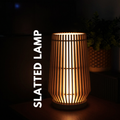 A glowing slatted table lamp with vertical wooden slats, shown in a dim room setting with a plant in the background. White vertical text on the left reads 'SLATTED LAMP'.