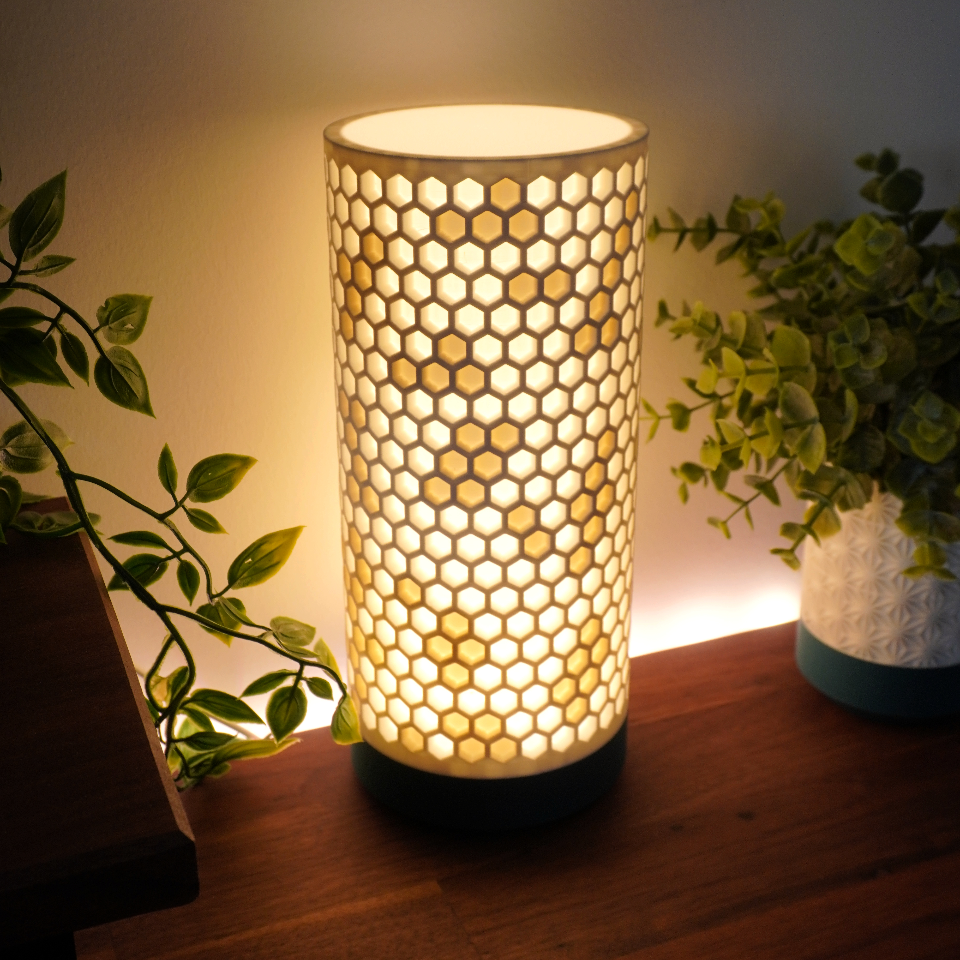 A cylindrical table lamp with a glowing honeycomb-patterned shade, placed on a wooden surface next to potted plants in a dimly lit room.