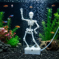 A white plastic skeleton fish keeper decoration standing on a base inside an aquarium, surrounded by green and pink artificial plants, small orange fish, bubbles, and a clear tubing connection.