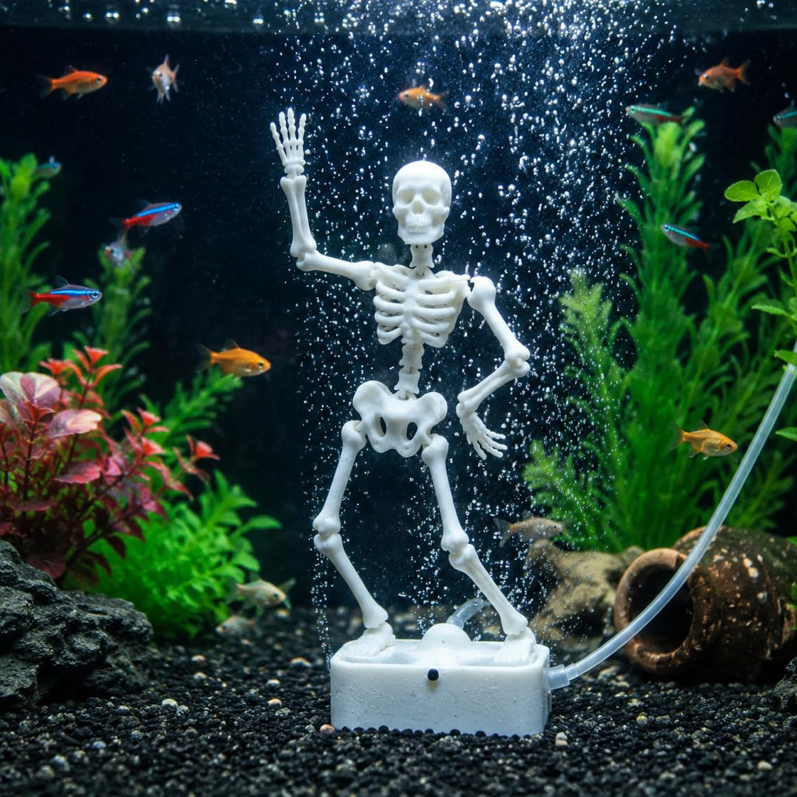 A white plastic skeleton fish keeper decoration standing on a base inside an aquarium, surrounded by green and pink artificial plants, small orange fish, bubbles, and a clear tubing connection.